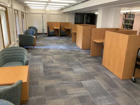 An open space with cubicles on the right. Pairs of lounge chairs bookending coffee tables on the left wall. Windows on left wall.
