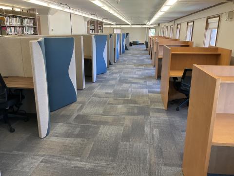 A corridor formed by carrels on the left against the wall and pods on the right. Bookstacks behind pods on the left, windows on right wall.