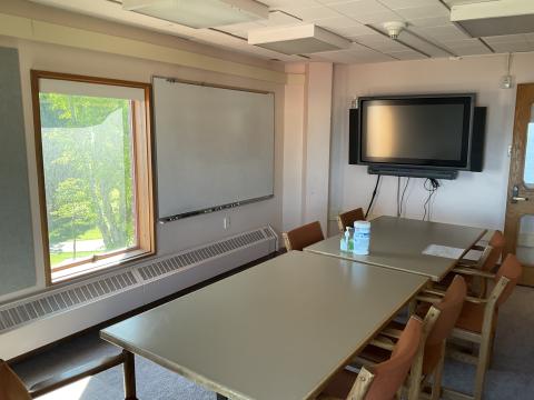 A room with a long table surrounded by chairs. Smartboard on back wall, whiteboard and window on left wall. Doorway in right corner.