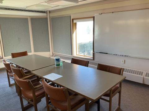 A room with a long table surrounded by chairs. Whiteboard on back right. Window centered in far wall.