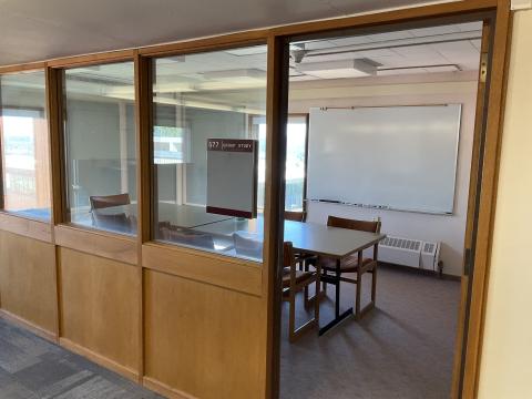 A room with a long table surrounded by chairs viewed through doorway. Windowed wall adjacent to doorway. Whiteboard on back right. Window centered in far wall.