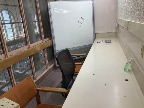 A small narrow room with a counter on the right wall. Chairs lined at counter. Whiteboard in back of room. Windows into Reading Room form left wall.