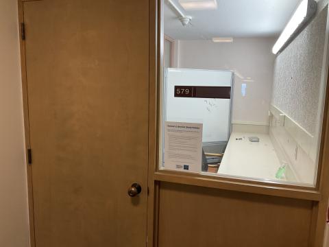 A small narrow room with a counter on the right wall, viewed through window adjacent to doorway.. Chairs lined at counter. Whiteboard in back of room.
