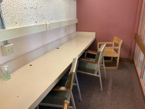 A small, narrow room with a counter on the left wall. Three chairs are lined up at the counter. Right wall is a window into the Reading Room.