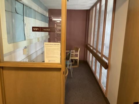 A small, narrow room, viewed through doorway, with counter with chairs on left wall. Windowed wall adjacent to doorway. Right wall is window into Reading Room.