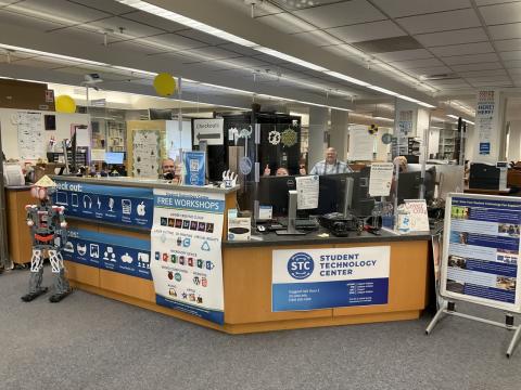 Large Service desk with signs showing items to checkout, Free workshops, Student Technology Center, and 3 people behind the desk and a robot in front. 