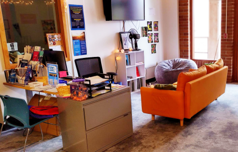 A room with a cluttered desk covered in papers and pamphlets. Orange couch and beanbag perpendicular. String lights. Warm lighting with cozy atmosphere.