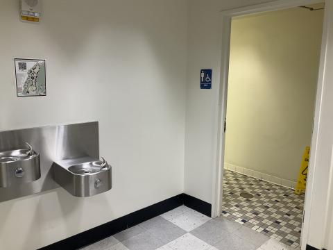 A doorway into a men's restroom on the right. Tiled flooring. Water fountains on the wall to the left.