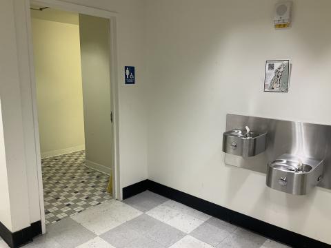 An open door into a women's restroom on the left. Water fountains on the wall to the right. Tiled flooring.