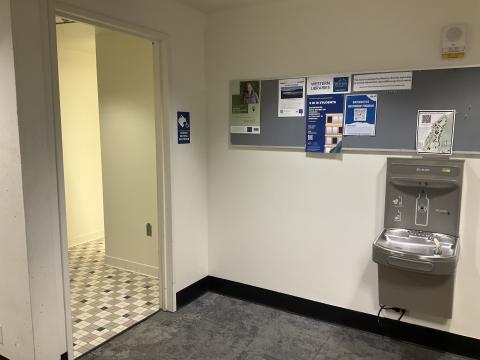 Open door into a gender-neutral bathroom on the right. Water-bottle filling fountain on adjacent wall to right. Pin board with posters above fountain.