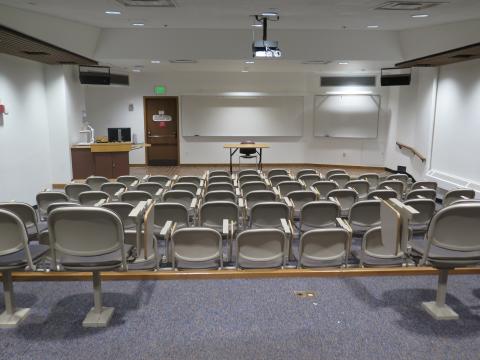 Lecture hall with tiered seating, looking toward the front where there is a whiteboard, podium, and rows of grey seats in front