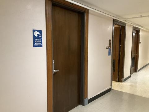 A closed door for a gender-neutral restroom in hallway. Signage about locations of gender-neutral restrooms left of door. Two more doors down hallway.