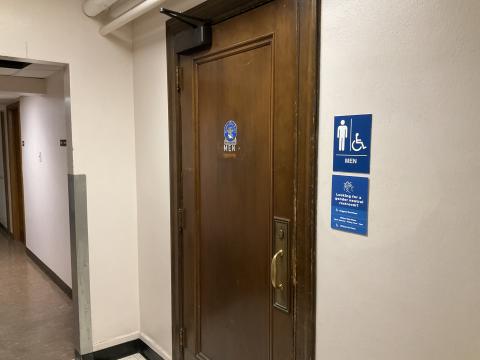 A closed door for a men's restroom. Signage to right of door with locations of gender-neutral restrooms. Open doorway to left.
