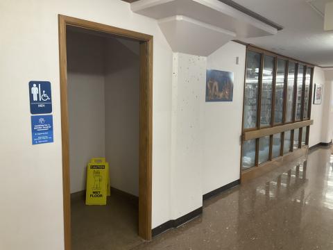 Open door to men's restroom. Signage denoting gender to left of door. Windows along adjacent wall to right.