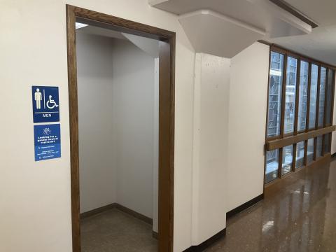Open door to men's restroom. Signage denoting gender to left of door. Windows on adjacent wall to right.