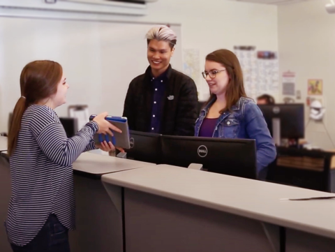 Student shows a device to two help desk staff standing across a tall counter; additional help desk staff member in the back behind a computer