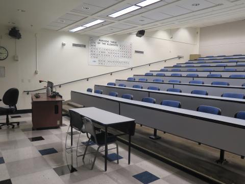 lecture hall with tiered seating, blue seats, periodic table on the wall.