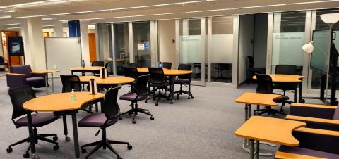 Room with crescent shaped tables surrounded by purple wheeled chairs. Whiteboard on back left. Glass doors to group study rooms along back wall.