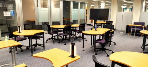 Room with crescent shaped tables surrounded by purple wheeled chairs. TV screen on back right. Glass doors to group study rooms along back wall. Table tents.