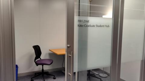 Small room seen through an open doorway. Long table with purple rolling chairs. Frosted glass label on door; HH 216E Kitto Graduate Student Hub.